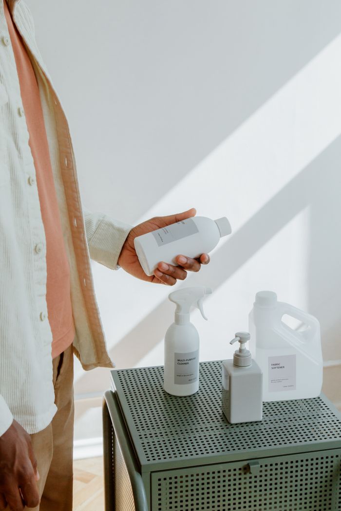 A man holds a plastic cleaning bottle while standing by a sunlit table with various products.